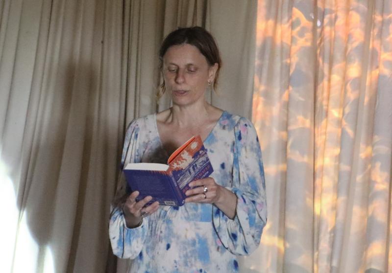 En person i mönstrad klänning står inomhus och läser i en bok, med ljusmönster projicerade på en gardin i bakgrunden.