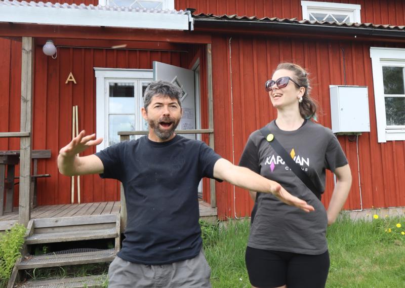 En man gestikulerar livligt medan en kvinna med solglasögon ler bredvid honom, båda står framför ett rött trähus med veranda.