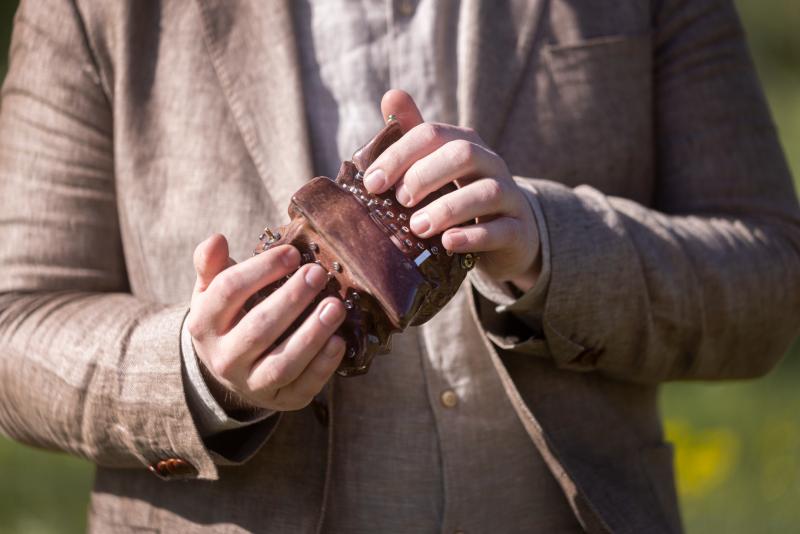 En person i beige kostymkavaj håller i och spelar på ett litet brunt dragspelsliknande musikinstrument utomhus.