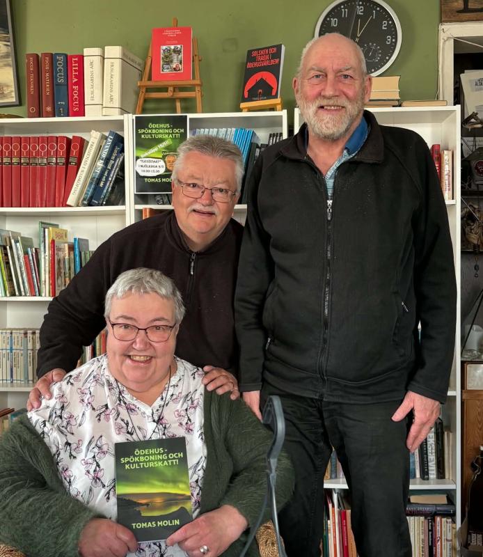 Tre äldre vuxna poserar tillsammans i ett rum med bokhyllor; den sittande kvinnan håller i en bok med titeln 