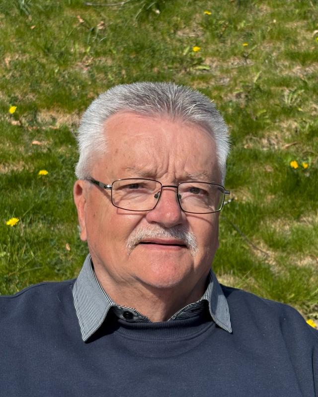 En äldre man med grått hår, glasögon och mustasch sitter utomhus på ett grässtrå med gula blommor, klädd i en mörk tröja över en skjorta med krage.