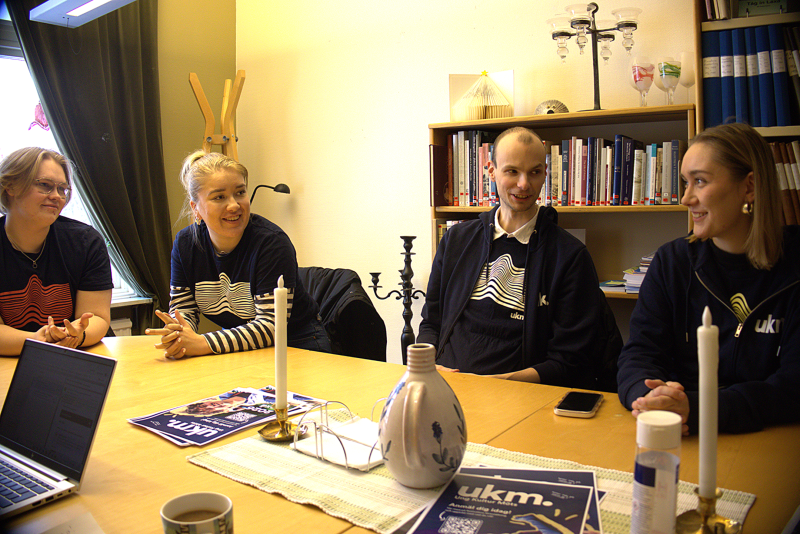 Fyra personer sitter runt ett bord på ett kontor och pratar och ler. På bordet står papper, stearinljus, en vas och muggar och i bakgrunden finns hyllor med böcker.