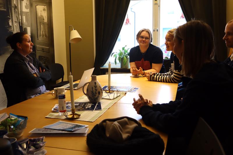 Fem personer sitter runt ett träbord i ett varmt upplyst rum och samtalar. Papper, tekanna och ljus står på bordet och dagsljuset faller in genom fönstret.