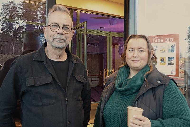 Två personer står framför en entré till en byggnad, den ena håller i en pappersmugg. En filmaffisch syns på väggen bredvid dem.