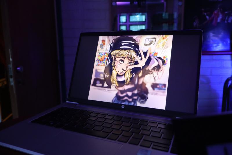 En bärbar dator visar ett digitalt konstverk i anime-stil av en blond karaktär som gör ett fredstecken inomhus, med ett svagt upplyst rum i bakgrunden.