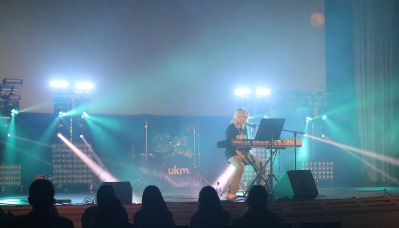 En musiker spelar keyboard och sjunger på en scen i starkt ljus, med trummor och andra instrument i bakgrunden och en publik i silhuett.