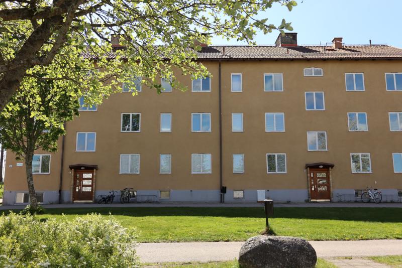 Ett fyra våningar högt, brunt hyreshus med många fönster, två entrédörrar och flera cyklar parkerade utanför, beläget bredvid en grön gräsmatta och träd en solig dag.