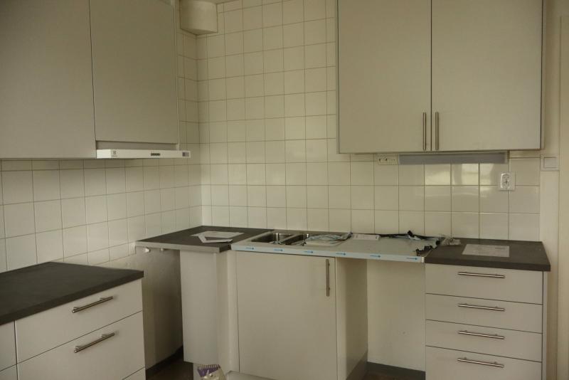 Ett modernt, oavslutat kök med vita skåp, grå bänkskivor, kaklat stänkskydd och några papper och förpackningar på bänken.