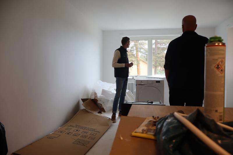Två personer står i ett sparsamt möblerat rum med tvättmaskin och torktumlare vid fönstret, omgivna av ouppackade kartonger och renoveringsmaterial.