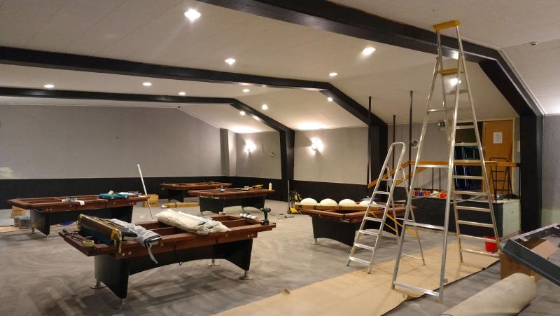 Ett rum med flera nedmonterade biljardbord, verktyg, stegar och utrustning, vilket tyder på pågående renoverings- eller installationsarbete. Taklamporna är tända och golvet är delvis täckt.