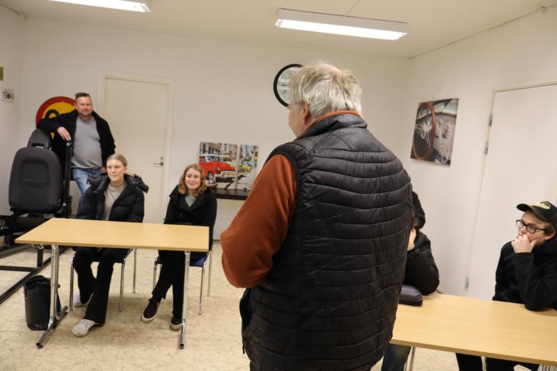 En man står och pratar med en grupp sittande människor i en klassrumsmiljö med bord och bilbilder på väggarna.