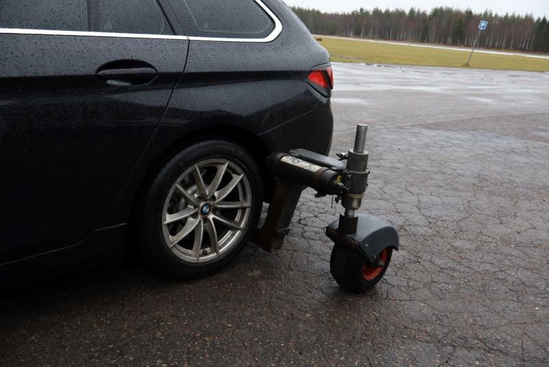 En svart bil med en hjulklämma fäst på det vänstra bakdäcket, parkerad på en våt, sprucken trottoar.