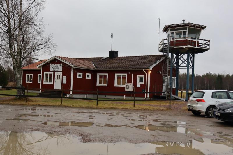 En röd byggnad och ett högt utsiktstorn ligger i ett lerigt område med parkerade bilar i närheten. Ett bladlöst träd står till vänster. Mulen himmel i bakgrunden.