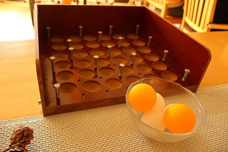 En spelplan i trä med vertikala metallpinnar ligger på ett bord bredvid en glasskål med två orange och två vita pingisbollar.