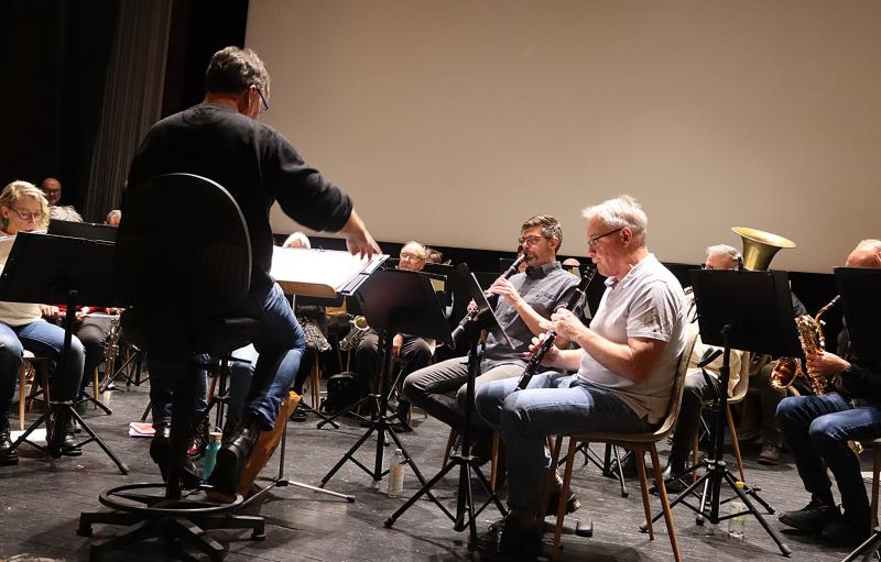 En dirigent leder en orkesterrepetition; musikerna spelar olika instrument sittande på scenen med notställ framför sig.