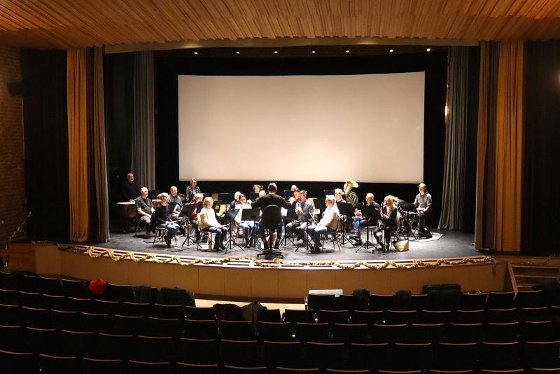 En dirigent leder en sittande orkesterrepetition på en scen framför en tom teater med en storbildsskärm i bakgrunden.