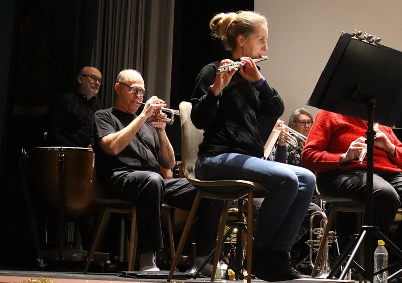 En grupp musiker sitter på scenen och spelar blåsinstrument och slagverk; en kvinna i förgrunden spelar flöjt, medan andra spelar trumpeter och timpani i bakgrunden.
