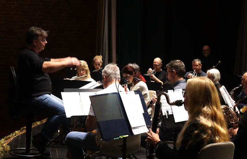 En dirigent leder en sittande orkester under en repetition, medan musikerna läser noter och spelar olika blåsinstrument på en svagt upplyst scen.