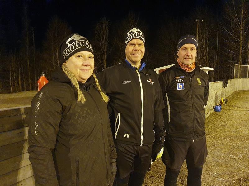 Tre vuxna i vintersportkläder och mössor står utomhus på en idrottsplats på natten, med träd och ett trästaket i bakgrunden.