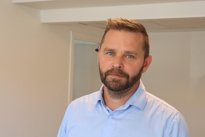 Man med kort skägg och ljusbrunt hår, klädd i ljusblå skjorta med krage, står inomhus med ett neutralt ansiktsuttryck.