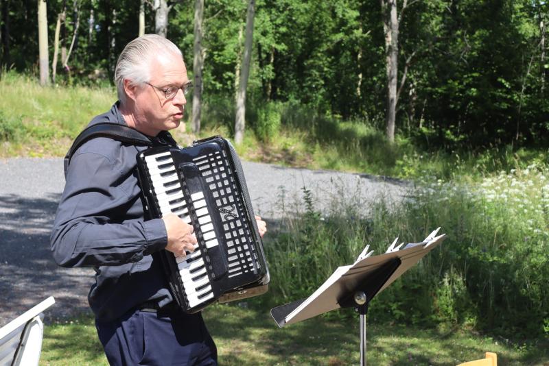 En man med glasögon spelar dragspel utomhus, stående bredvid ett notstativ med noter, omgiven av grönska och solljus.