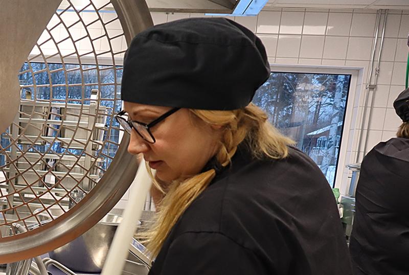 En kvinna med glasögon och svart kockmössa arbetar i ett storkök nära stor utrustning och ett fönster som visar en snöig utomhusscen.