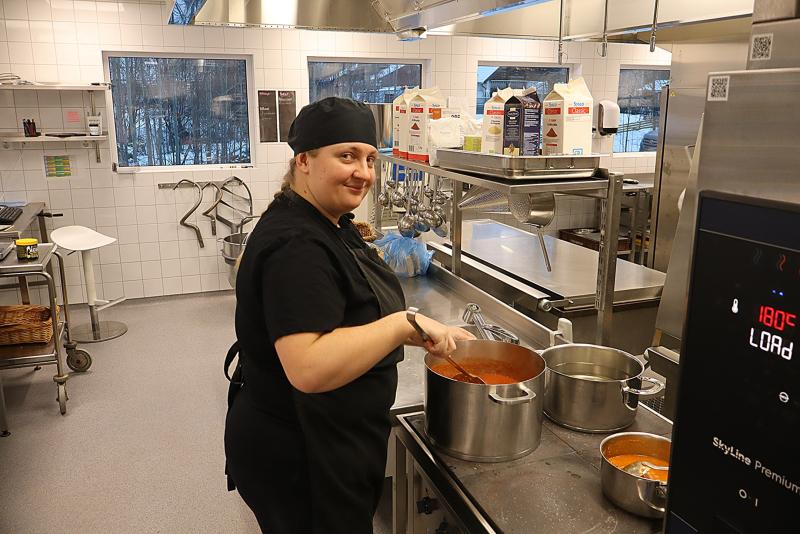 En person i svart kockuniform rör om i en kastrull med sås i ett storkök med rostfria apparater och påsar med ingredienser på en bänk.