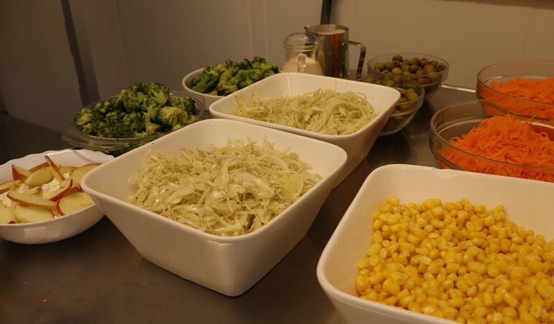 En mängd olika färska salladsingredienser, inklusive majs, strimlade morötter, oliver, broccoli, äppelskivor och strimlad kål, visas i skålar på en disk i rostfritt stål.