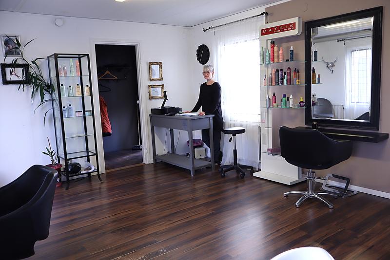 En kvinna står bakom ett skrivbord i en modern frisörsalong med trägolv, stylingstolar och hyllor fyllda med hårprodukter.