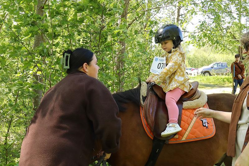 En ung flicka med hjälm sitter på en häst med orange sadel medan två vuxna hjälper henne i ett skogsområde.