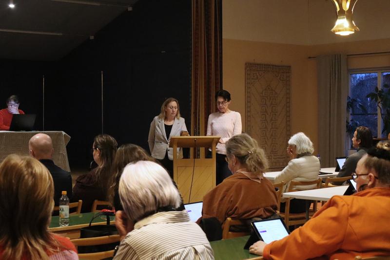 Två kvinnor står på ett podium och talar till en publik som sitter vid bord med bärbara datorer i ett konferensrum. Andra personer ses arbeta vid ett bord och på en bärbar dator i bakgrunden.