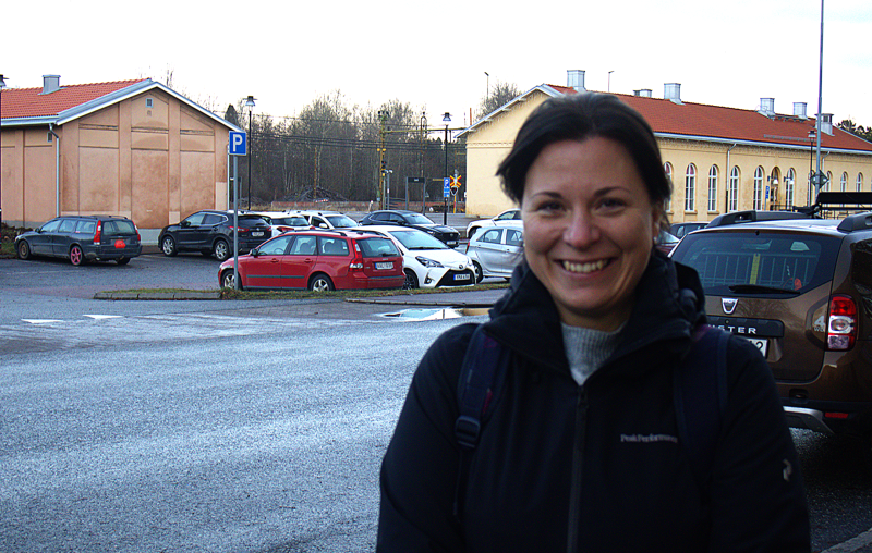En kvinna i svart jacka står och ler på en parkeringsplats med flera parkerade bilar och gula byggnader i bakgrunden.