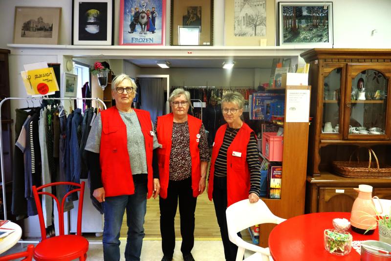 Tre kvinnor i röda västar står inne i en secondhandbutik, omgivna av klädställningar, hyllor med föremål och inramade konstverk på väggarna.