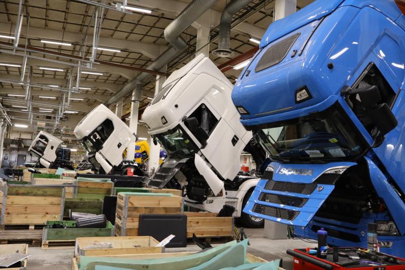 Fyra lastbilshytter, i vitt och blått, lutar framåt i fabriksmiljö, omgivna av trälådor och industriutrustning.