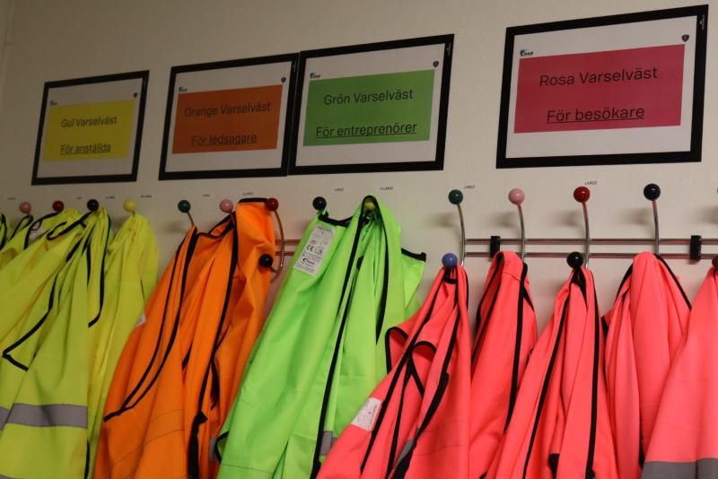 Västar med hög synlighet i gult, orange, grönt och rosa hänger på krokar under motsvarande skyltar på svenska som anger deras avsedda bärare.