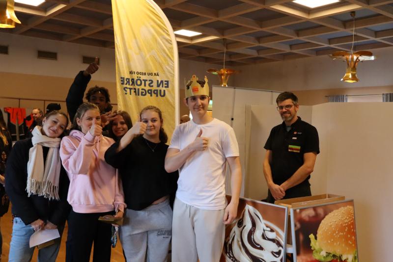 En grupp människor poserar för ett foto inomhus; en person bär en krona och en annan person gör en tummen upp. En man står bakom ett bord med en hamburgerdisplay.