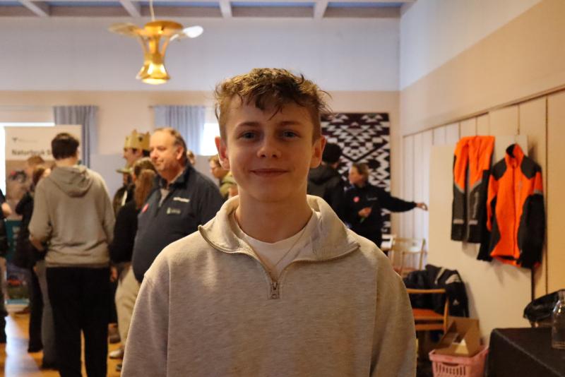 Ung person i ett rum med andra i bakgrunden. Rummet har taklampa och rockar hängande på väggen.