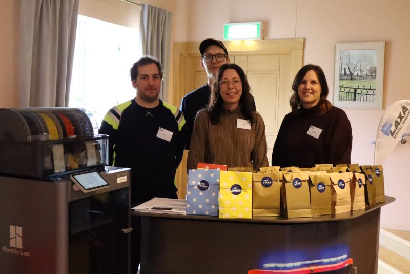 Fyra personer står bakom ett bord med påsar kaffe och en kaffemaskin till vänster, i ett rum med ljusa väggar och konst på väggarna.