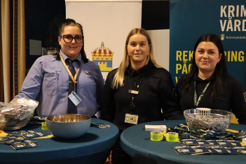 Tre personer står bakom bord med broschyrer, skålar och reklamartiklar vid ett evenemang. De har uniformer och linor på sig. Banderoller i bakgrunden visar händelseinformation.