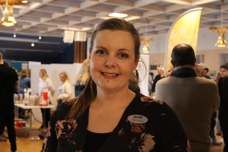 En kvinna som ler vid ett evenemang, klädd i en blommig topp och namnskylt, med människor i bakgrunden i ett väl upplyst rum.