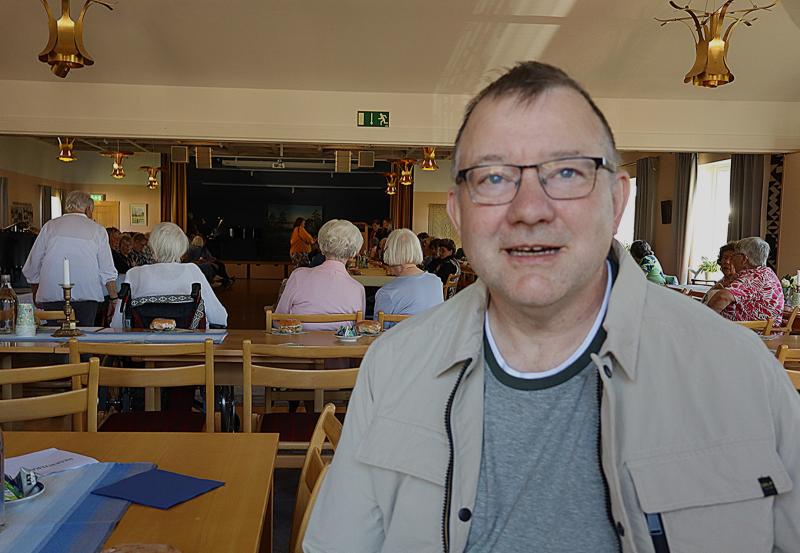 En man med glasögon sitter vid ett bord i ett väl upplyst rum med flera andra personer sittande i bakgrunden, vända mot en scen eller ett presentationsområde.