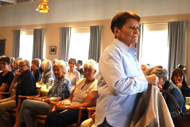 En kvinna står och håller i en kappa i ett rum fyllt med sittande äldre personer, alla vända framåt, med naturligt ljusinsläpp genom fönster med gardiner.