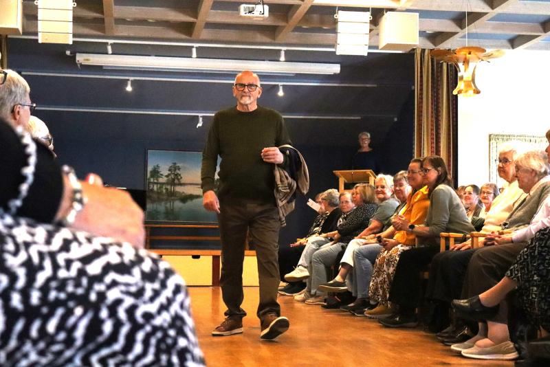 En äldre man går på en gångbana i ett rum med sittande människor på båda sidor, medan en kvinna står vid ett podium i bakgrunden.