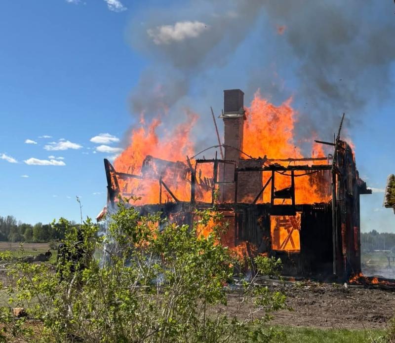 Ett trähus är uppslukat av lågor och rök stiger upp mot himlen. Det omgivande området består av grönska och ett öppet fält.
