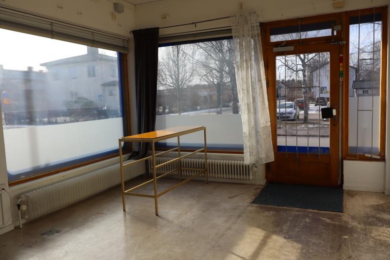 Tomt rum med stora fönster, ett litet träbord och utsikt över träd och byggnader utanför. Dörr med glasruta leder till utsidan.