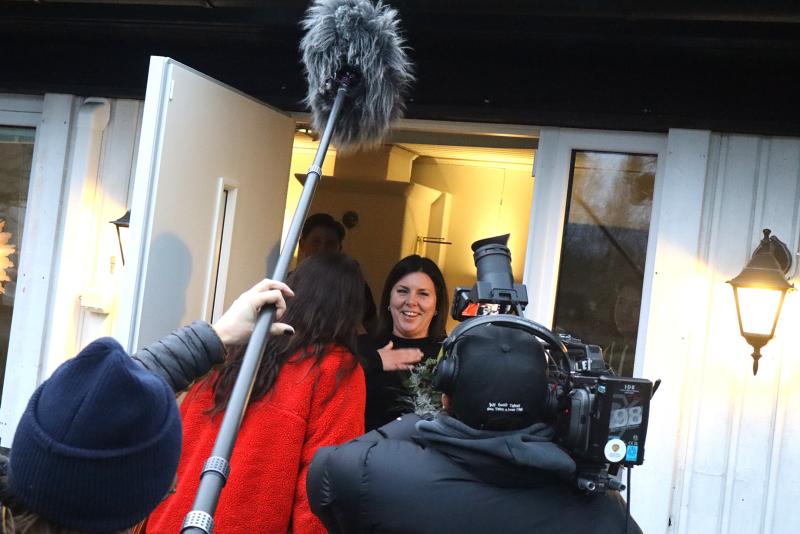 En kvinna står i en dörröppning och ler mot ett kamerateam som filmar henne med en bommikrofon och videokamera; en annan person i röd rock står mitt emot henne.