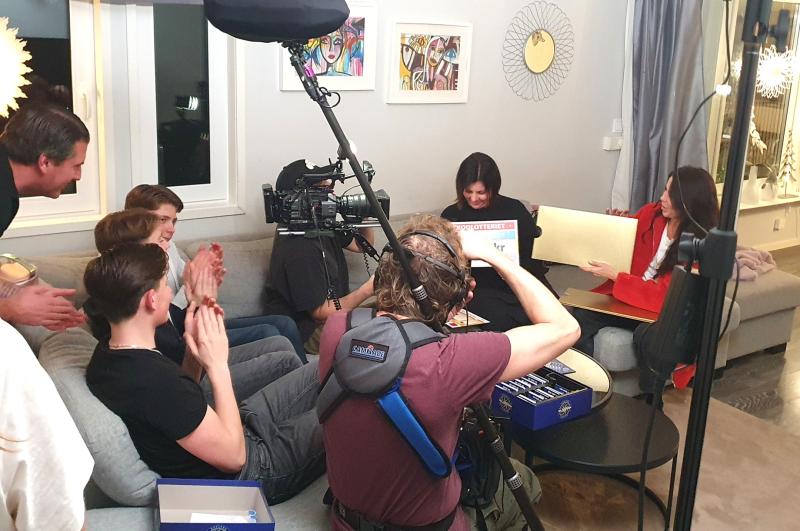 Ett kamerateam filmar en grupp människor som sitter i en soffa, klappar och ler medan en kvinna håller upp en brädspelslåda i en vardagsrumsmiljö.