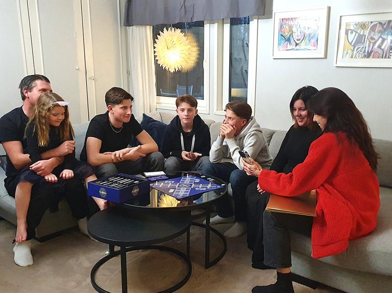 Sju personer i olika åldrar sitter i en soffa i vardagsrummet, engagerade i samtal och brädspel, med spelpjäser och ett bräde synliga på bordet framför dem.