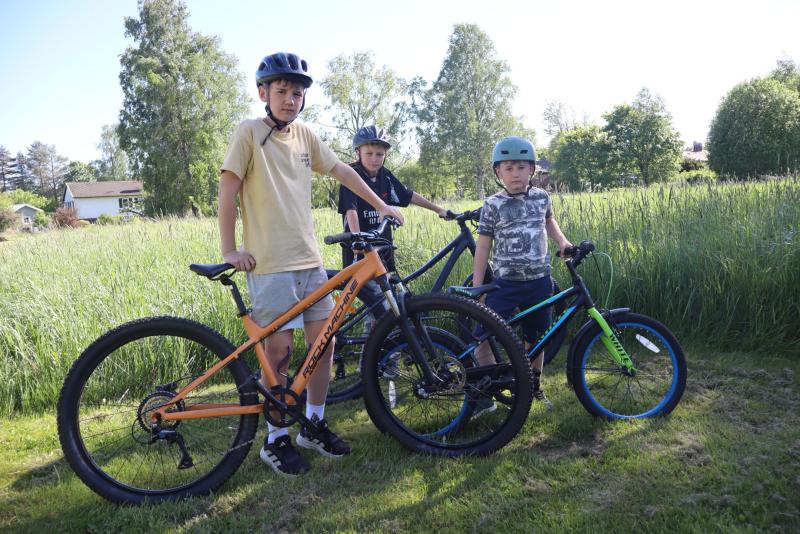 Tre pojkar med hjälmar står utomhus med sina cyklar på en gräsyta, träd och buskar syns i bakgrunden en solig dag.