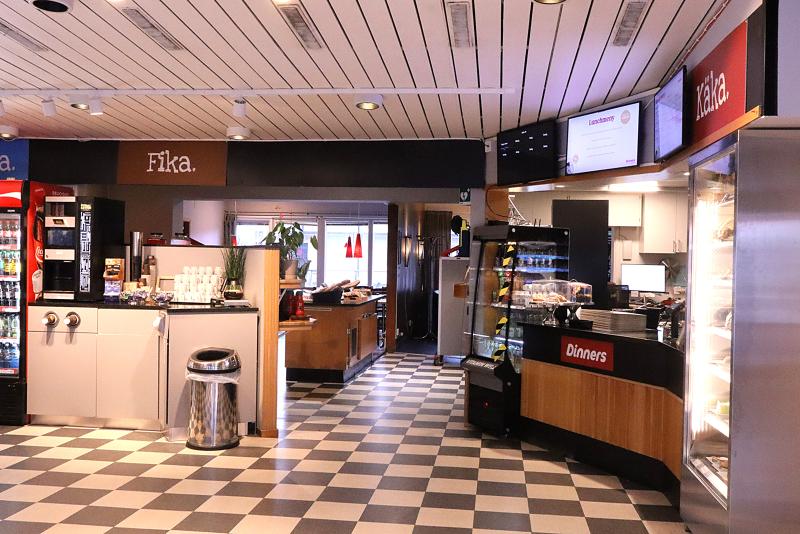 Cafeteria med rutigt golv, dryckesmaskiner, matdiskar, en papperskorg och skyltar för 